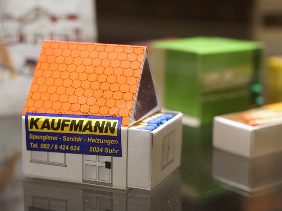 Eine Zündholzschachtel in Form eines Hauses mit Giebeldach ist zu sehen mit der Aufschrift «Kaufmann: Spenglerei, Sanitär, Heizungen».