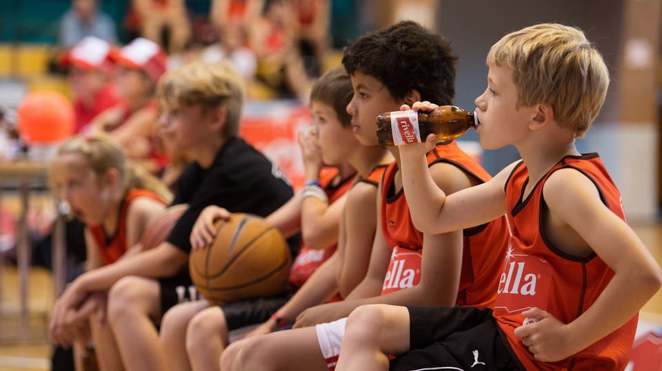 Kinder mit Rivella-Shirts trinken am Rande eines Basketballspiels Rivella. 