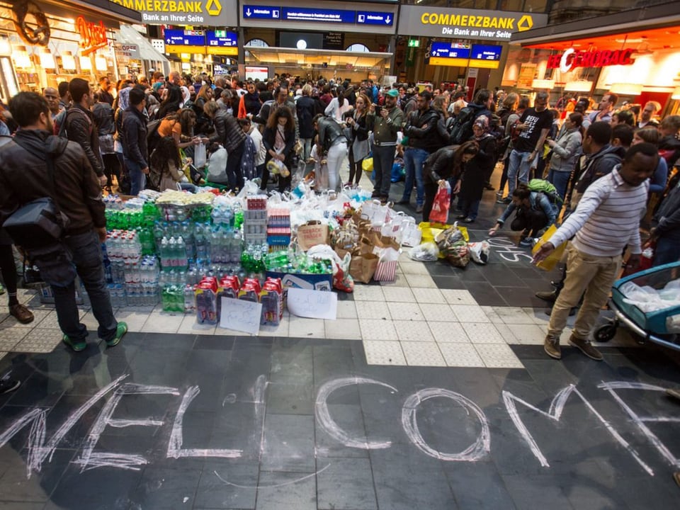 Bahnhofshalle in Frankfurt, auf dem Boden steht Welcome, im Hintergrund Wasser und Essen aufgestapelt. 