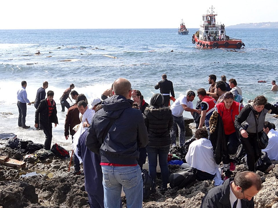 Zahlreiche Menschen an einem Strand, im Wasser treiben Teile des Schiffwracks