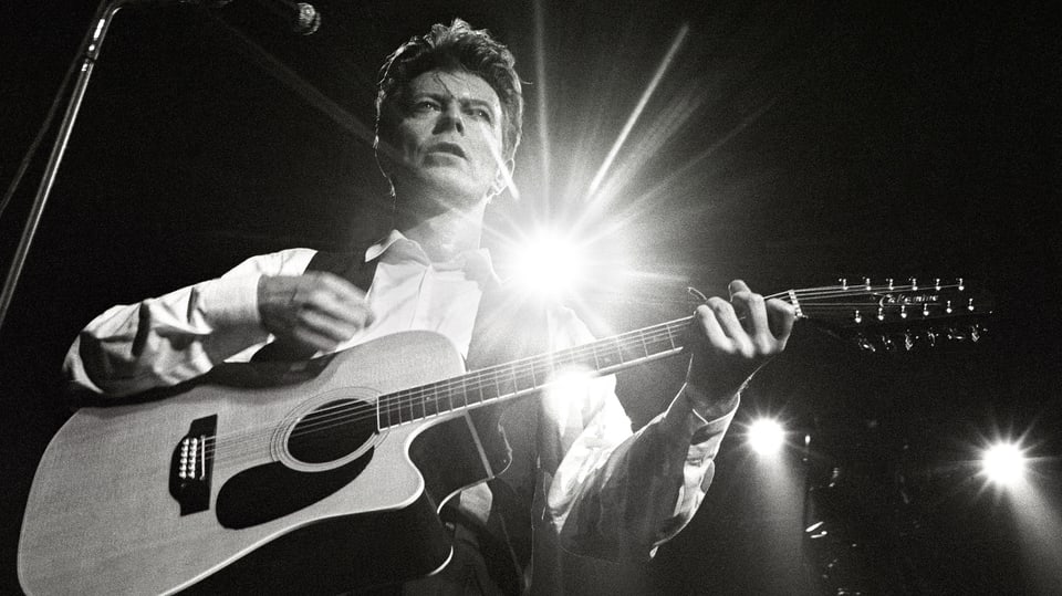 David Bowie posiert auf der Bühne mit Gitarre.