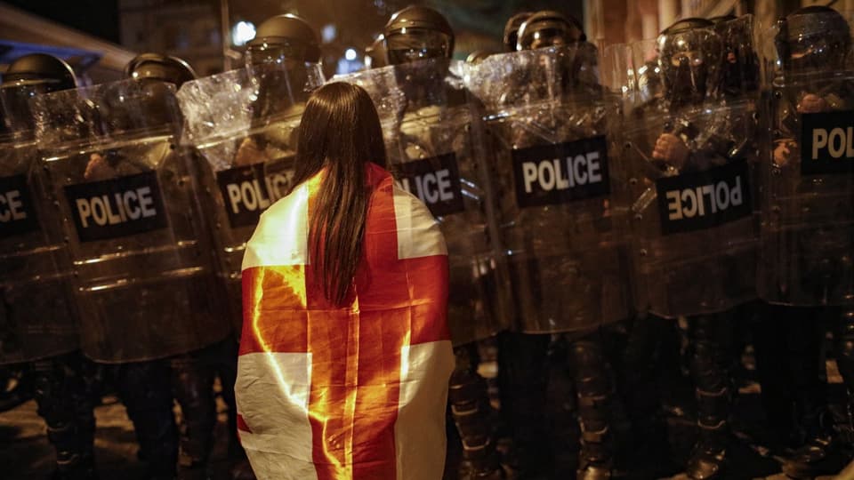 Frau mit Flagge steht vor einer Reihe Polizeibeamter in Schutzkleidung bei Nacht.