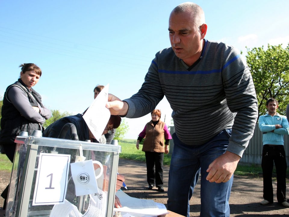 Eine gläserne Wahlurne ist am Rande einer Strasse aufgestellt. Ein Mann wirft seinen Zettel ein.