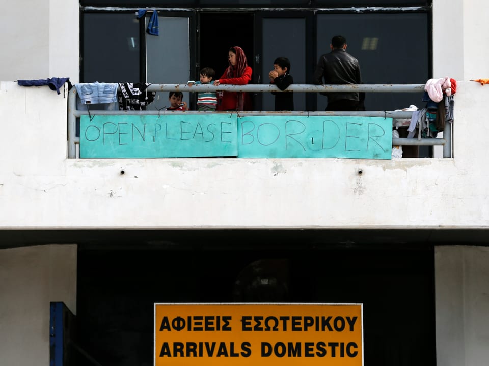 Flüchtlinge stehen auf einem Balkon, an dem ein Protest-Banner hängt mit der Aufschrift: "Open pelase border."