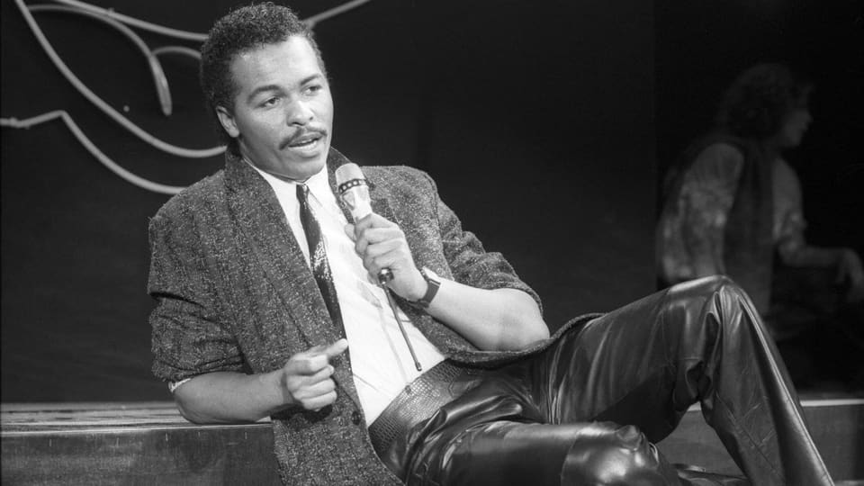 Ein schwarzer Mann sitzt mit einem Mic in der Hand auf einer Bühne.