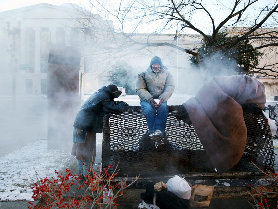 Obdachlose wärmen sich an einem dampfenden Entlüftungsschacht.