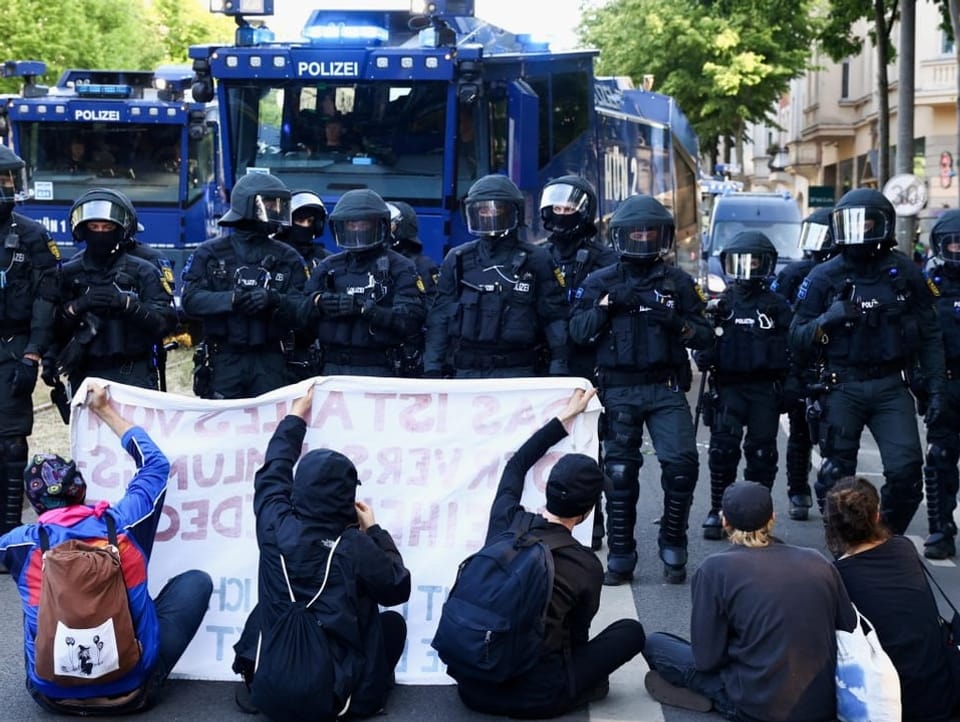 Mehrere Personen sitzen vor einer Polizeiblockade. Sie halten ein Transparent hoch.