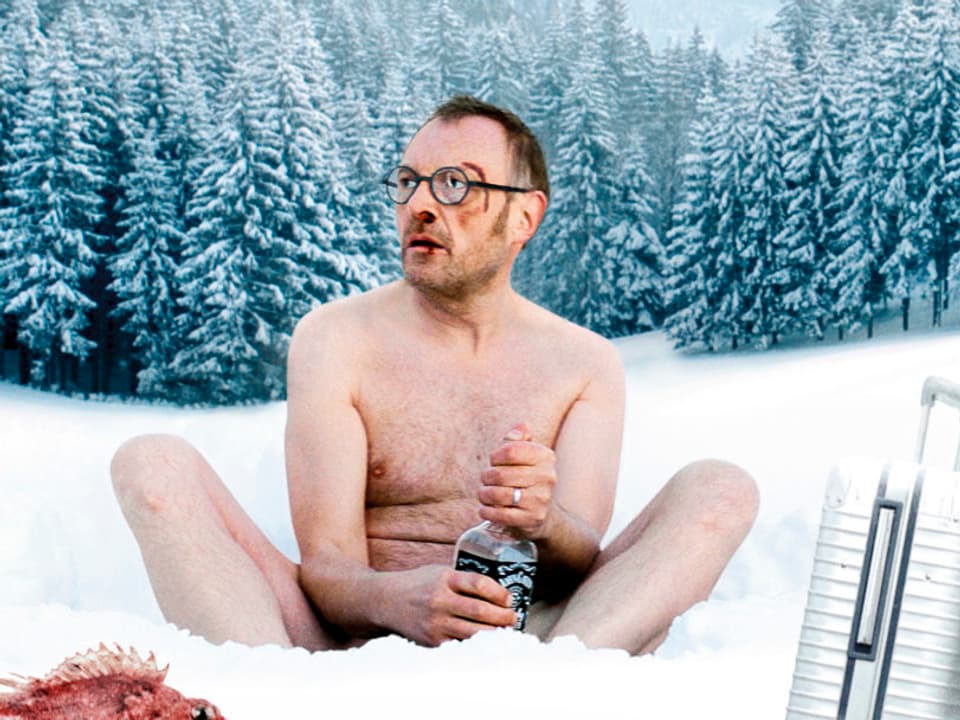 Josef Hader sitzt halbnackt im Schnee mit einer Flasche Whiskey, einem Fisch und einem Koffer.