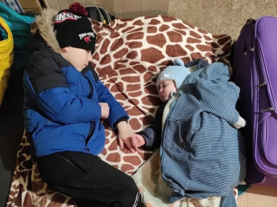 Ein Baby und ein Junge in Winterklamotten schlafen nebeneinander in einem Keller. 