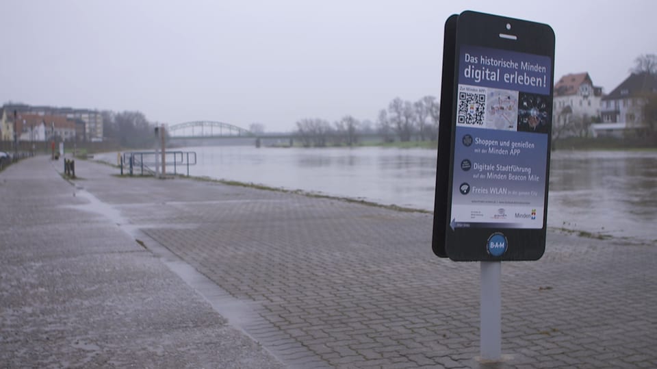 Ein Smartphone so gross wie ein Mensch am Flussufer von Monden. Es wirbt für die «Beacon-Mile».