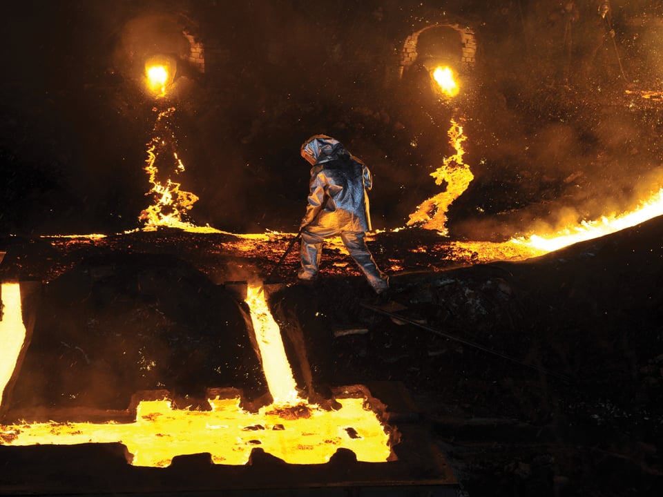 Ein Mann in Schutzkleidung rührt mit einem Stab in einer lava-artigen, glühenden Masse.