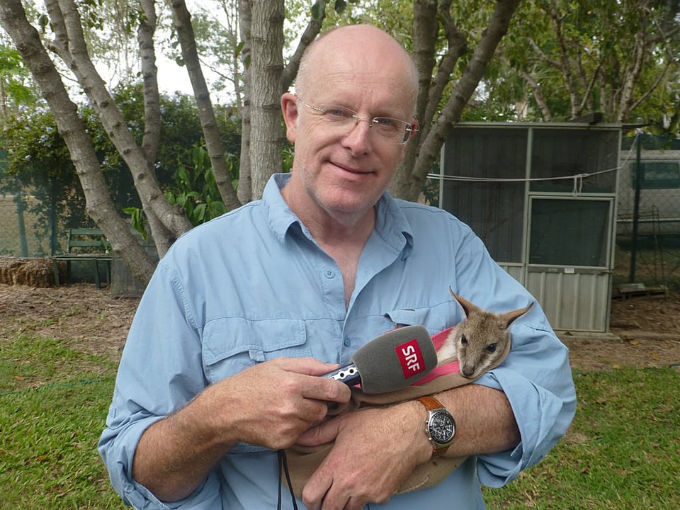Urs Wälterlin hält ein kleines Känguru in den Armen und streckt ihm ein Mikrofon ins Gesicht.