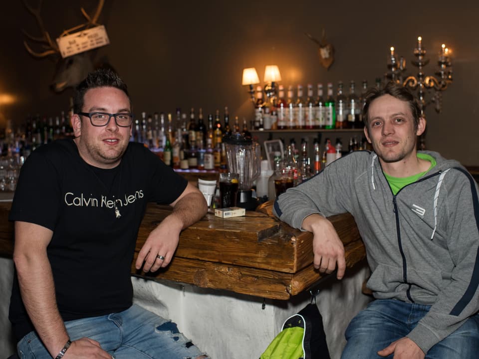 Zwei Männer sitzen an einer Bar und schauen in die Kamera. Der Mann links trägt ein schwarzes T-Shirt und eine Brille, der andere einen grauen Kapuzenpullover über einem grünen T-Shirt.