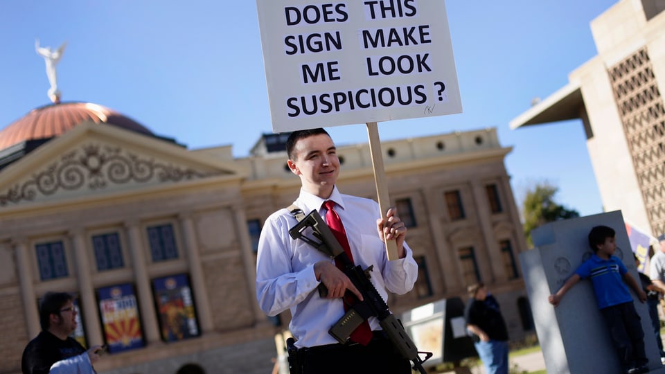 Ein Mann mit einem Gewehr hält ein Transparent hoch: «Macht mich dieses Plakat verdächtig?»