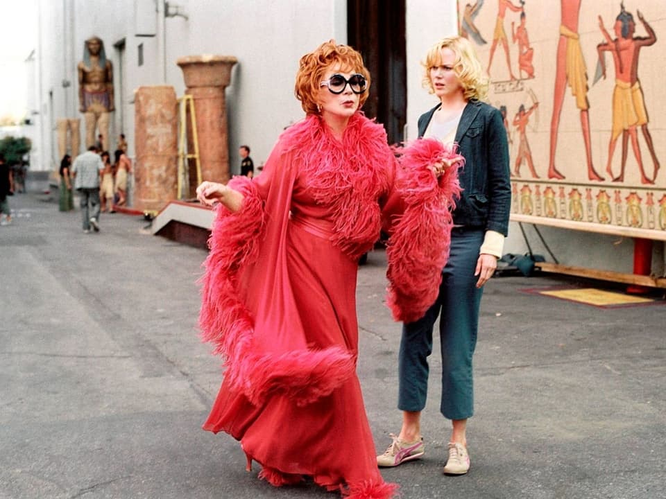 Zwei Frauen in einem Filmset, eine in extravagantem roten Federoutfit, die andere in lässiger Kleidung.