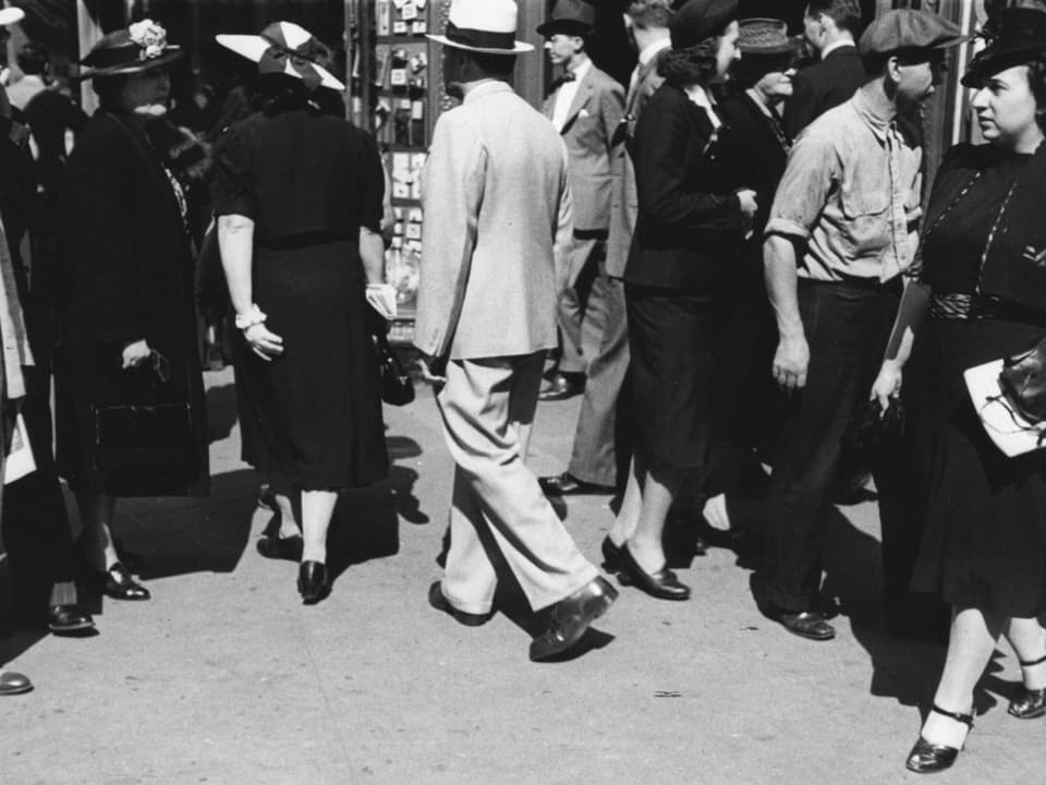 Auf einem schwarz-weissen Foto ist ein Bürgersteig mit vielen Menschen drauf zu sehen.