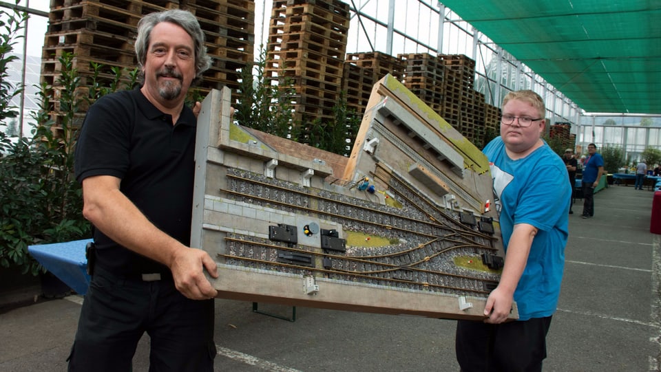 Zwei Menschen transportieren einen Teil einer Gartenbahn