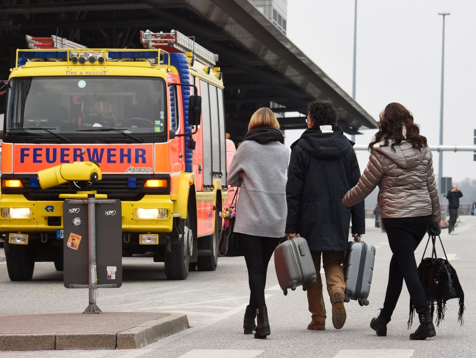 Menschen mit Koffern vor dem Hamburger Flughafen. Im Hintergrund Feuerwehr-Autos. 