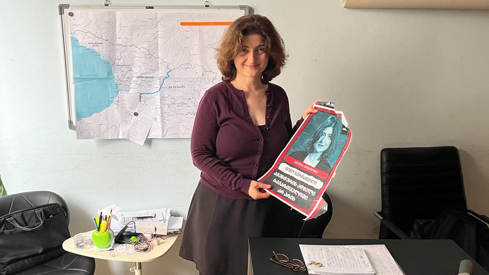 Eine Frau im Büro zeigt ein Plakat. Darauf ist ein Foto von ihr zu sehen.
