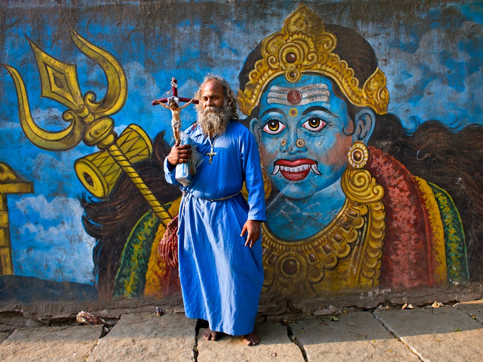 Ein Inder in einem blauen Kleid und einem Jesuskreuz. Hinter ihm ist eine Zeichnung auf einer Wand.