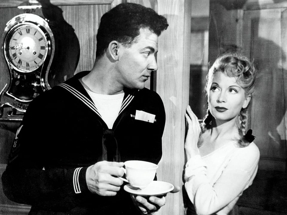 Ein Mann in Matrosenuniform mit einer Kaffeetasse in der Hand blickt auf eine Frau mit blondem, zu Zöpfen geflochtenem Haar.