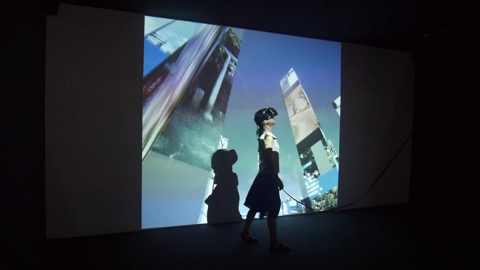 Eine Frau mit VR-Brille steht in einem dunklen Raum. Auf ihren Körper und an die Wand wird ein Bild projiziert, auf dem Wolkenkratzer zu sehen sind. 