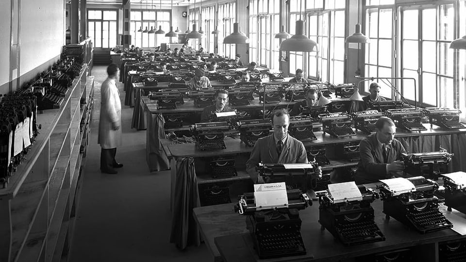 Schwarzweissfoto: In einem Fabrikraum sitzen an langen Bänken Menschen hinter Schreibmaschinen