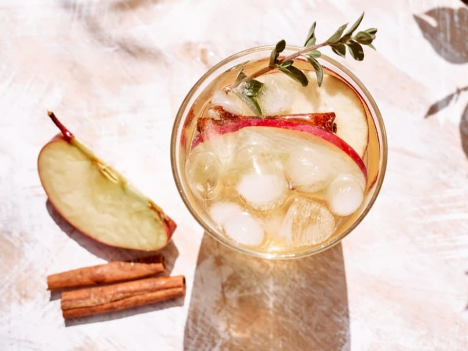 Cocktail mit Eis und Apfelschnitzen, daneben Zimtstangen