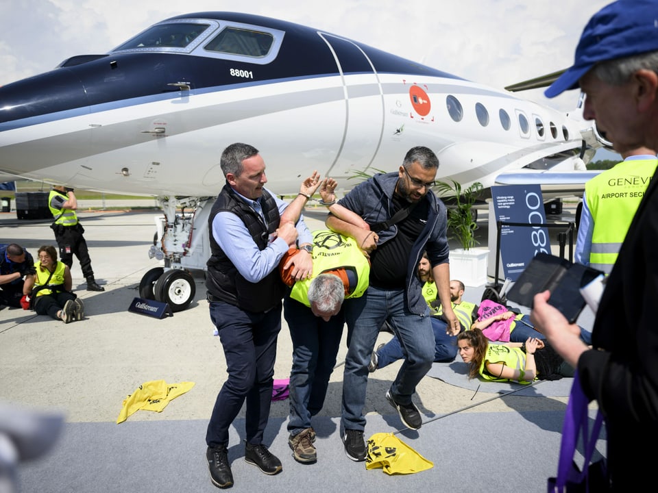 Mann wird mit zurückgehaltenen Händen von zwei Männern abgeführt. Im Hintergrund steht ein Flugzeug.
