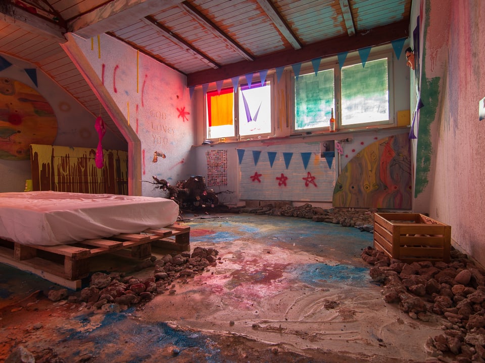 Zimmer mit einem Bett, grossen Steinen und vielen Farben an Wänden und auf dem Boden und dem Bett.