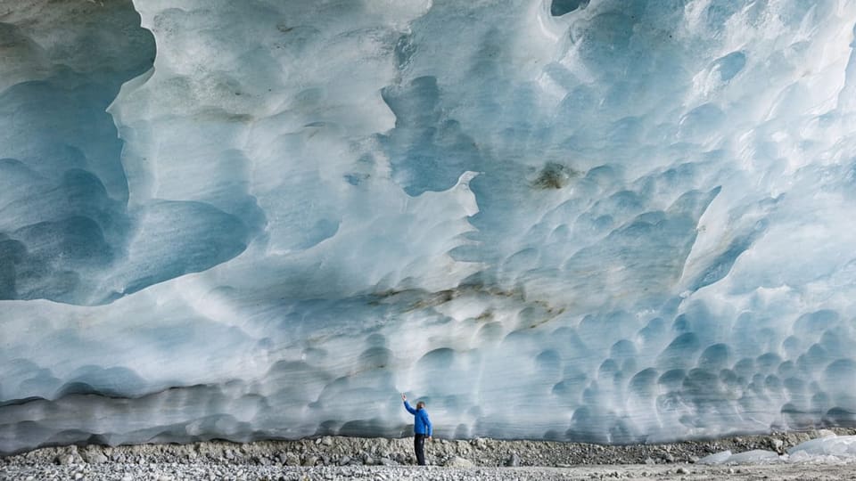Ein gewaltiger Eisblock eines Gletschers, darunter ein winziger Mann zu sehen.