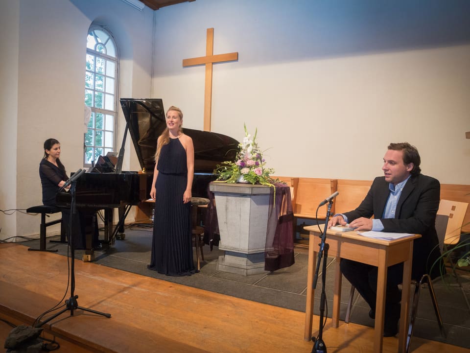 Eine Frau singt in einer Kirche, sie wird begleitet von einem Sprecher und einer Pianistin.