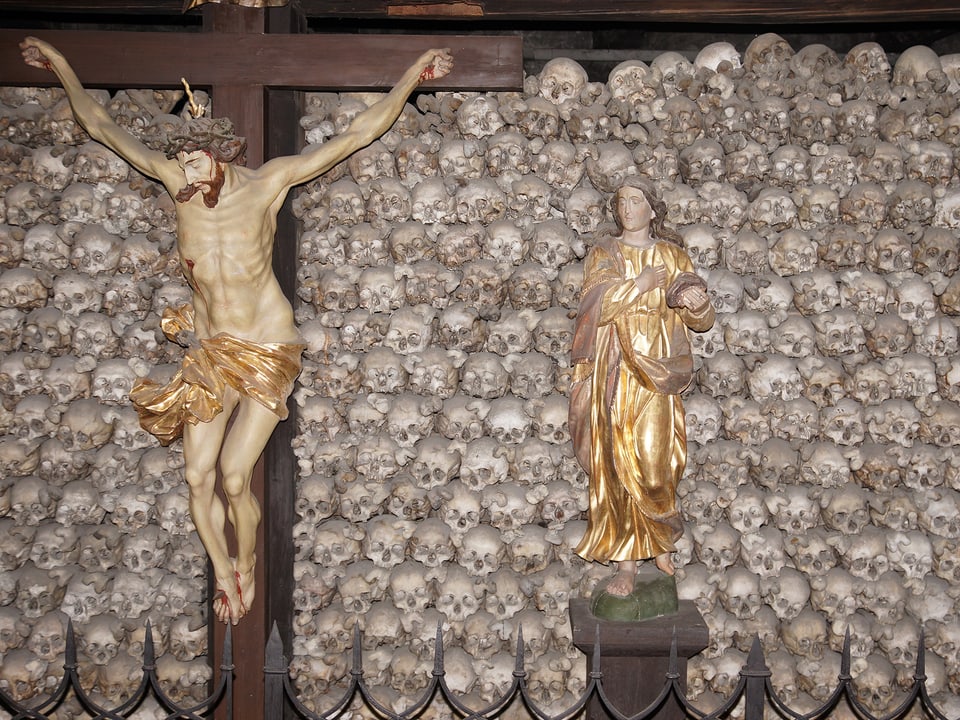 Ein Kruzifix und eine Marienstatue vor vielen aufeinander gestapelten Knochenschädeln.