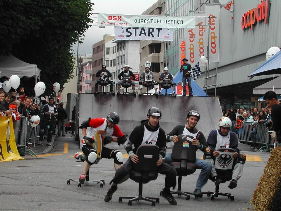 Bürostuhlrennen in Olten. 4 Personen sitzen mit Helmen auf Stühlen und fahren. Oben zu sehen: Die Rampe mit 4 weiteren Teilnehmern.