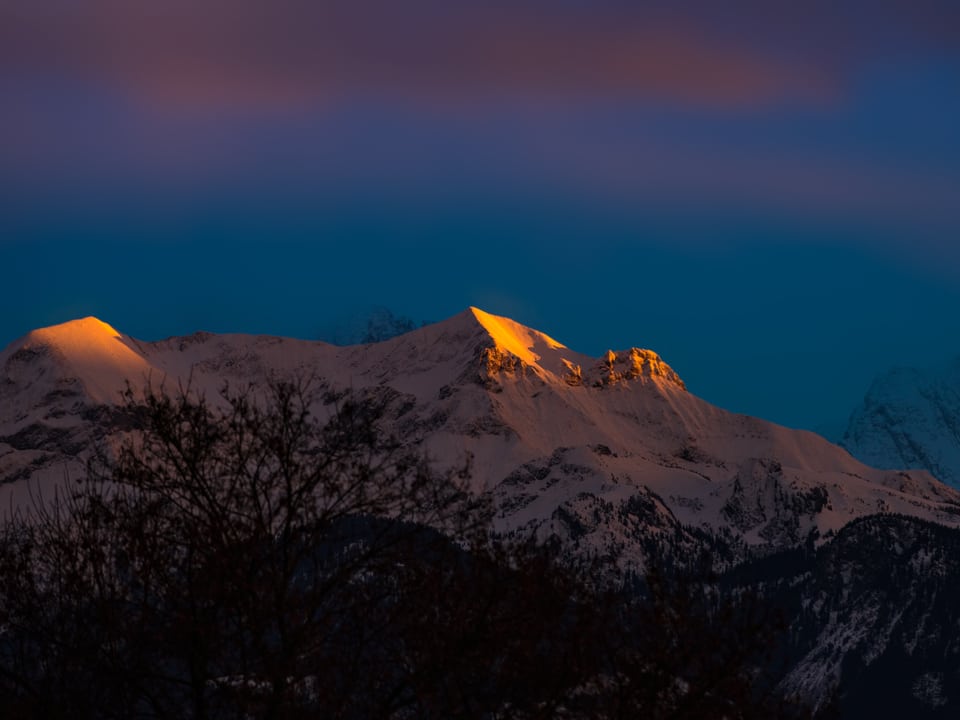 Auf der gegenüberliegenden Talseite ist eine verschneite Bergkette zu sehen. Die Gipfel erstrahlen in leuchtendem Gelb. Sie werden von der Sonne angeleuchtet, während es sonst darum herum grau ist.