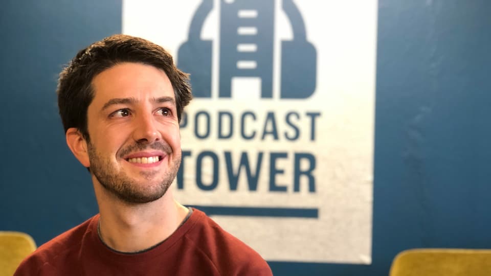 Ein Mann mit Dreitagebart und kurzen braunen Haaren sitzt vor einer Wand mit der grossen Aufschrift: Podcast Tower.