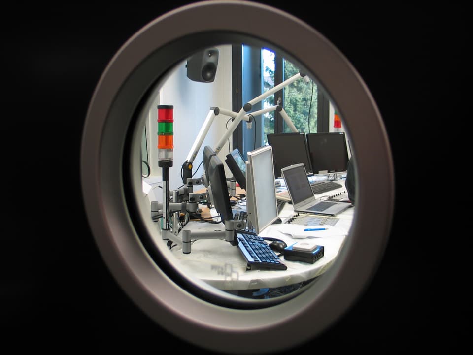 Blick durch ein rundes Fenster in ein Sendestudio.
