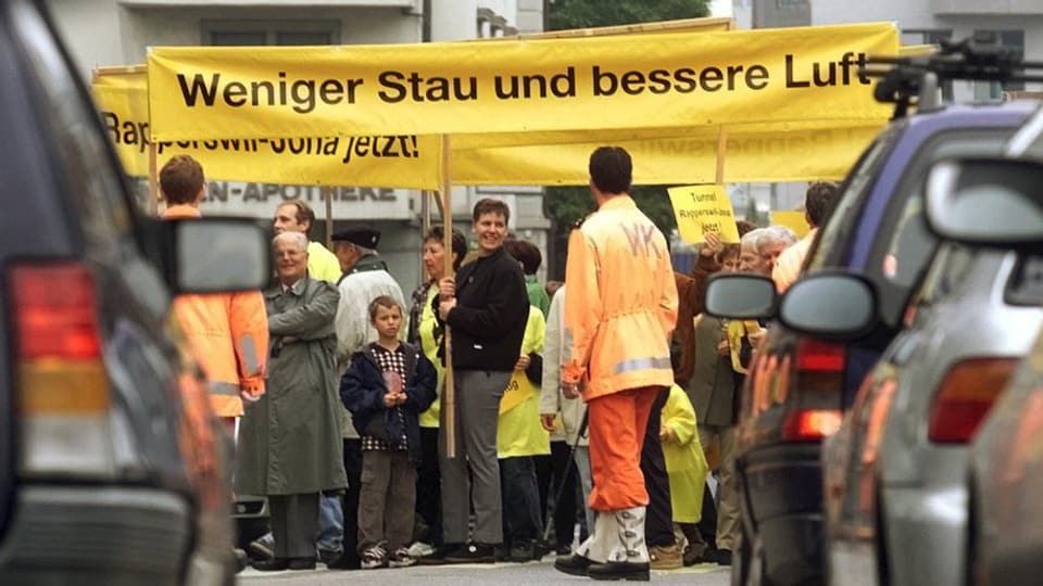 Zwischen zwei Autoreihen protestieren Menschen mit Bannern «Weniger Stau und bessere Luft»