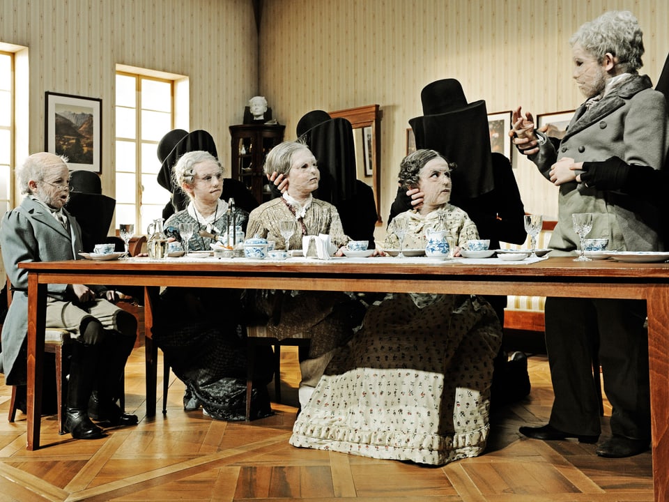 Als alte Menschen maskierte Kinder sitzen um einen Tisch, im Hintergrund schwarze Gestalten.