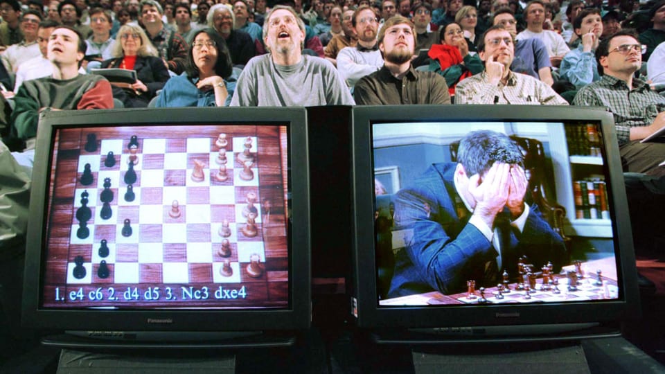 Auf zwei Monitoren sieht man den Schachcomputer und Kasparow vor einer sitzenden Menschenmenge.