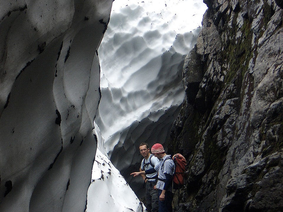 Zwei Wanderer stehen zwischen einer Mauer aus Schnee und einer Felswand.