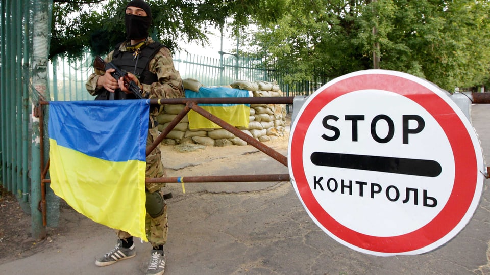 Ein Mann mit Gesichtsmaske steht vor einer Schranke auf der die ukrainische Flagge liegt.