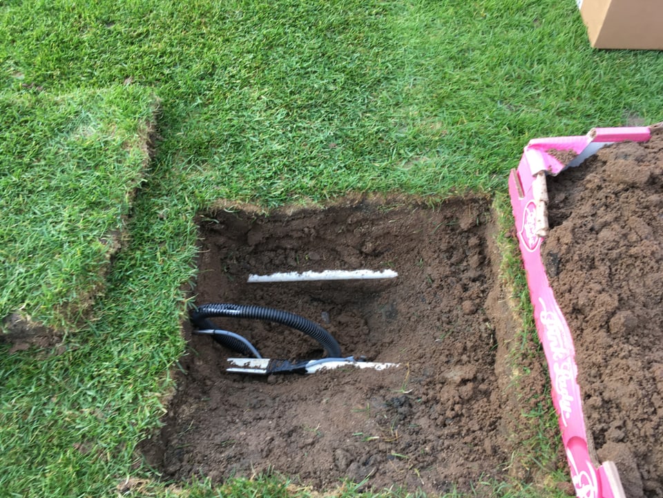 In den Rasen ist ein rechteckiges Loch gegraben. Im Loch werden eine Leitung und ein Themperaturfühler sichtbar. 