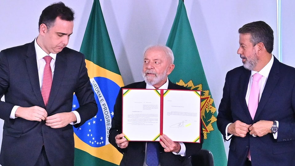 Brasiliens Staatspräsident Luiz Inácio Lula hält ein Dokument in die Luft, neben ihm stehen zwei Politiker.