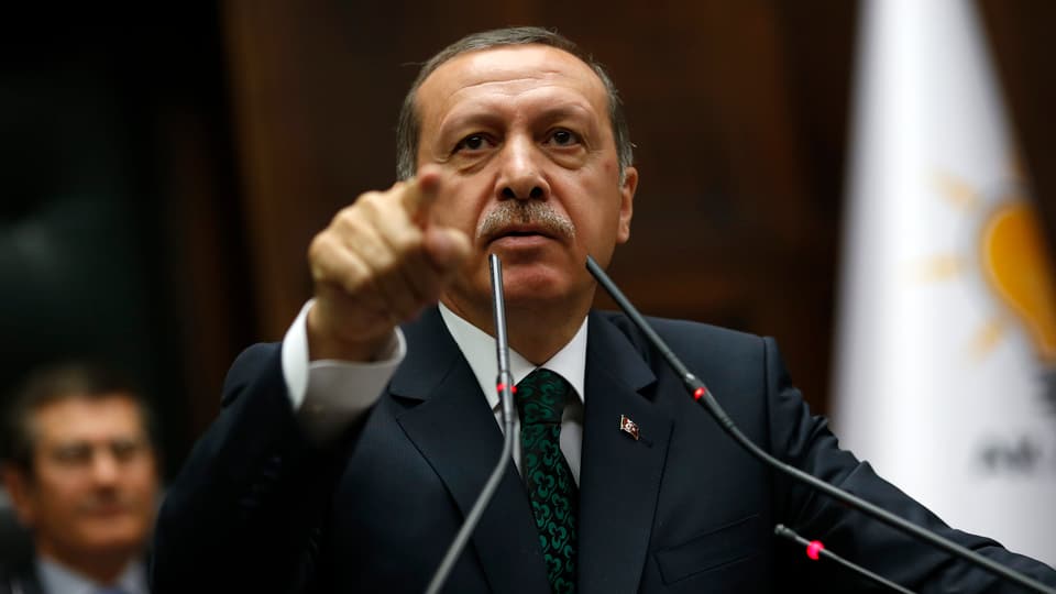 Erdogan vor zwei Mikrofonen, die rechte Hand zeigt ausgestreckt in Richtung Kamera.