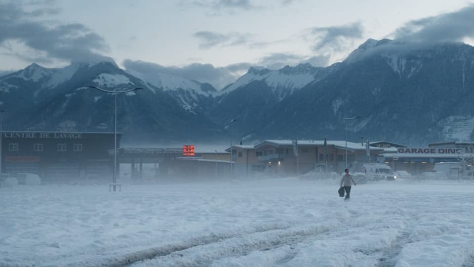 Foto einer Totalen: unten Schnee, hinten die Berge, davor trostlose Dörfer. eine Gestalt läuft im Schnee