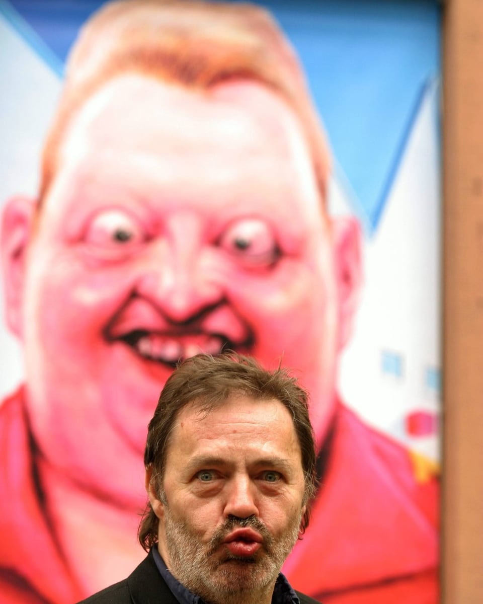 Mann mit dunkelgrauem Bart zieht Grimasse vor Bild von rosarotem Mann mit irrem Blick.