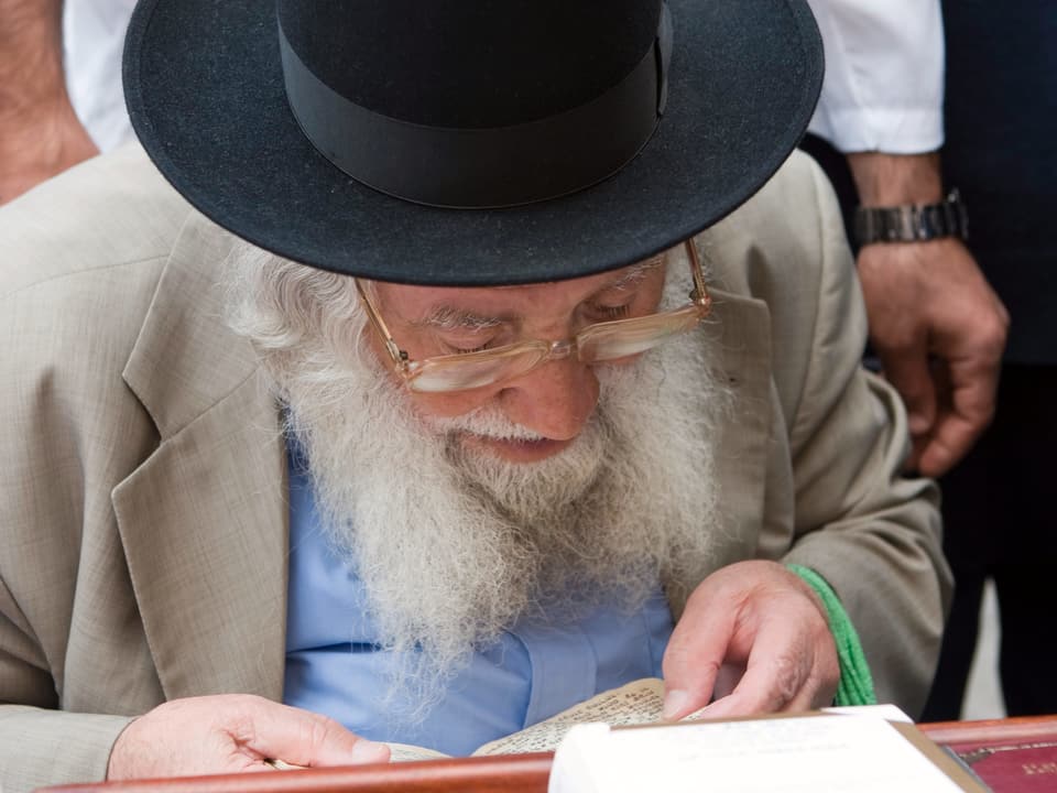 ein alter Mann mit Bart und Schwarzem Hut liesst mit einer goldenen Brille aus einem Judischen Gebetsbuch.