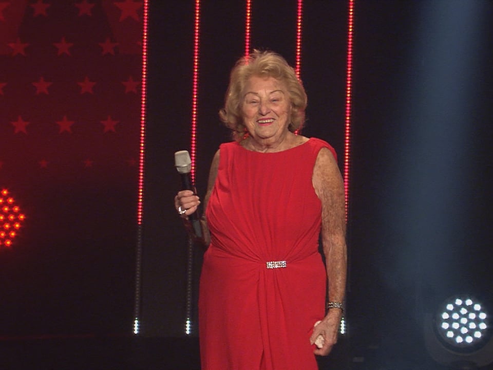 Eine ältere Frau in einem roten Kleid steht auf der Bühne und hält ein Mikrophon in der Hand.