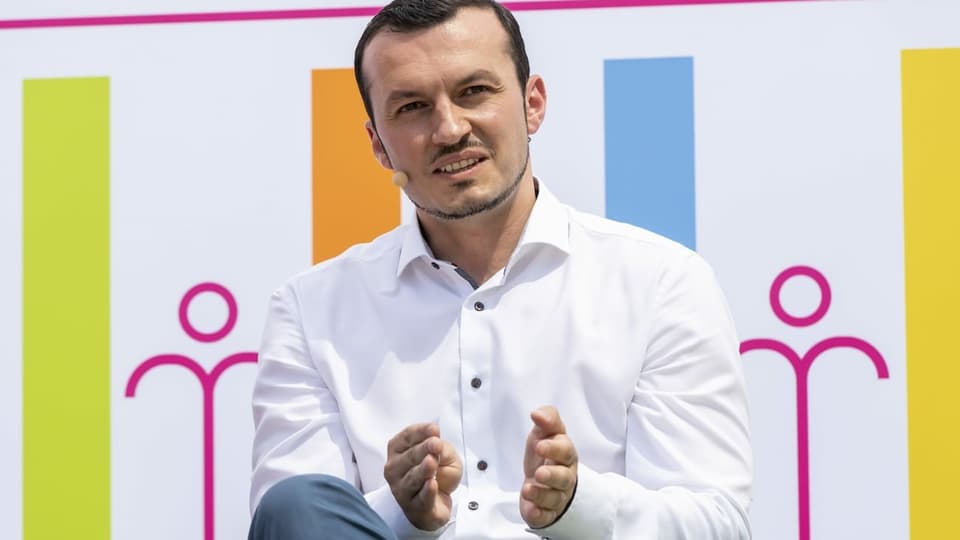 Ein Mann mit Mikrofon sitzt vor einer farbigen Plakatwand.